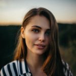 A person with long, brown hair wearing a striped shirt stands outdoors, the backdrop of a blurred natural landscape subtly capturing a hint of sunset. An aerial surveyor might use a UAV to capture such serene vistas. The individual looks directly at the camera with a neutral expression.