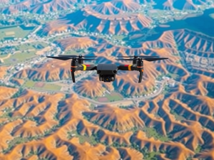 A drone with propellers is flying over a landscape characterized by rugged, orange-hued hills. The view shows a contrast between the barren hills and green patches with scattered buildings below. The scene captures a clear, sunny day.
