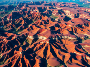 Aerial view of a vast desert landscape with vibrant red sand dunes and deep valleys, illuminated by sunlight. The undulating terrain creates a striking pattern of shadows and highlights, emphasizing the natural beauty and ruggedness of the region.
