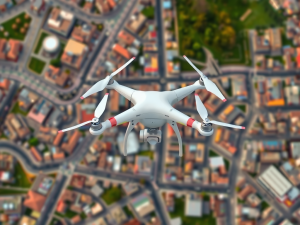 A white drone with red stripes is flying over an aerial view of a city. The cityscape below shows various buildings, roads, and green spaces, captured in a slightly blurred background, indicating the drone's movement.